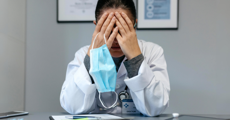 An exhausted doctor in a white lab coat sits with her head in her hands, visibly overwhelmed and suffering from compassion fatigue and burnout.