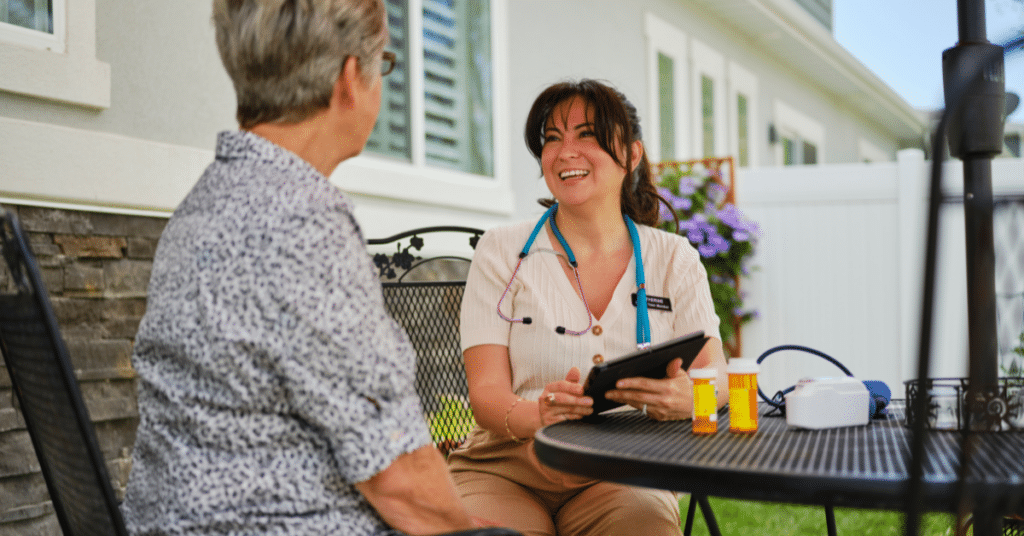 The image shows a woman healthcare provider and an elderly patient in a serene rural home setting. They are seated outdoors on a comfortable patio set, engaged in a warm conversation. The healthcare provider is smiling kindly, attentively listening and talking to the elderly patient, who radiates a sense of ease and contentment. The natural, peaceful backdrop of the rural environment enhances the sense of a personalized and compassionate healthcare experience. This scene encapsulates the reasons why healthcare jobs in rural areas are highly valued, highlighting the close, community-oriented relationships and the rewarding nature of providing care in such settings.